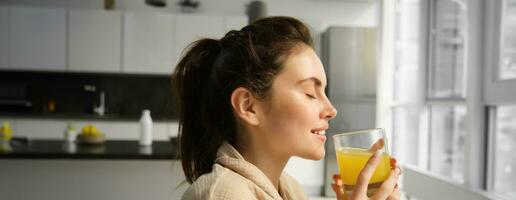 Close up portrait of beautiful woman enjoys her morning, drinks glass of orange juice with pleasure and delight, stands in living room in bathrobe photo