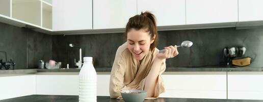 Portrait of young beautiful woman in bathrobe, eating cereals for breakfast, leans on kitchen worktop, looking at her morning meal photo