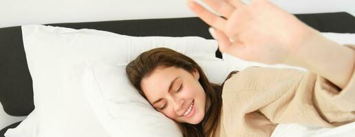 Portrait of gorgeous girlfriend stretching hand, blocking camera, lying in bed and resting on pillow, smiling while waking up from someone photo