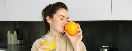 Close up portrait of young brunette, smells fresh orange, drinking juice from glass, enjoying healthy start of morning, wearing bathrobe photo