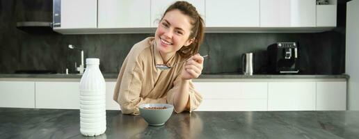 Laughing, beautiful woman in bathrobe, eats cereals with milk, holding spoon, smiling and looking happy, posing in kitchen and leaning on worktop photo