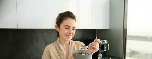 Close up portrait of smiling, beautiful woman eating cereals with milk, holding spoon and bowl, looking outside window in morning, standing near kitchen worktop photo