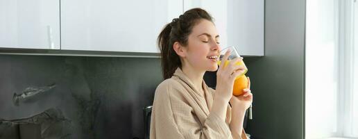 Portrait of happy woman in kitchen, wearing bathrobe, drinking orange juice, freshly squeezed drink, smiling and laughing, food and drink concept photo