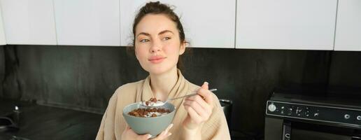 guapo morena mujer comiendo su desayuno, en pie en cocina cerca encimera y participación cuenco de cereales con leche, disfrutando su mañana, vistiendo acogedor bata de baño foto