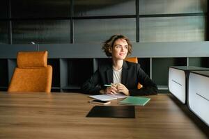 Young businesswoman waiting for start of meeting. Corporate woman in suit sitting in office, working on documents, waiting for client photo