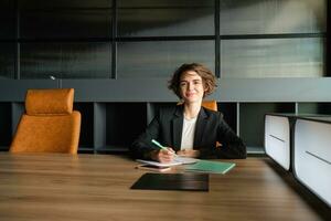 Young businesswoman waiting for start of meeting. Corporate woman in suit sitting in office, working on documents, waiting for client photo