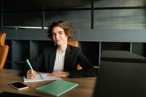 Portrait of successful brunette woman in suit, corporate manager in office, writing, preparing documents for meeting, reading case photo