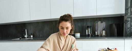 retrato de mujer escritura abajo lista de comestibles, haciendo notas en receta, sentado en cocina cerca verduras, preparando cena menú foto