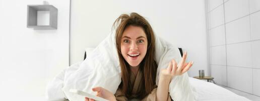 Impressed woman lying in bed and using mobile phone, looking amazed at camera by content online on smartphone photo