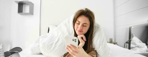 Portrait of beautiful brunette woman with smartphone, relaxing in her bed under blankets and smiling, checking social media feed on mobile phone app, wrap herself in duvet and enjoying lazy morning photo