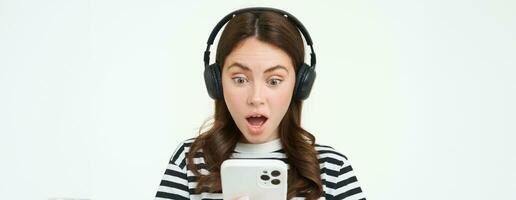 Portrait of young woman in wireless headphones, looks shocked at smartphone screen, reads message on mobile phone with dropped jaw, gasping from surprise, white background photo