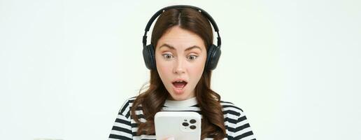 retrato de joven mujer en inalámbrico auriculares, mira conmocionado a teléfono inteligente pantalla, lee mensaje en móvil teléfono con caído mandíbula, jadeando desde sorpresa, blanco antecedentes foto