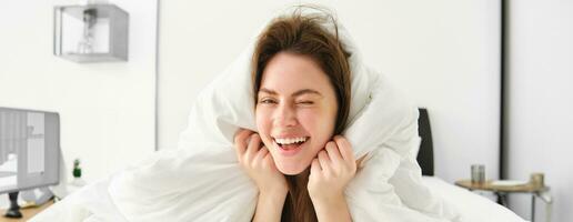 Portrait of gorgeous smiling woman, lying in bed covered in duvet, has messy hair, looking happy, relaxing in her bedroom, spending time in hotel room in morning photo