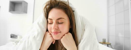 Cheerful woman feeling comfortable in bed, lying in bedroom covered in white sheets, smiling pleased, has messy hair in morning, waking up and looking happy photo