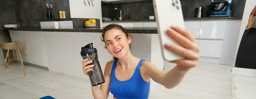 Portrait of sportswoman takes selfie with water bottle in living room, holds smartphone and poses for photo while doing fitness workout