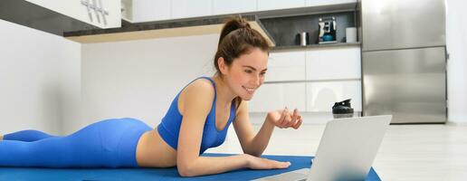 Image of young fitness girl talking online on laptop, video chats, laying on floor rubber mat, doing exercises, discuss workout training, yoga from home photo