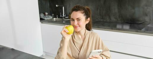 Image of attractive young woman in bathrobe, sits in kitchen, eats green apple and looks at mobile phone, uses smartphone app, orders food on application photo