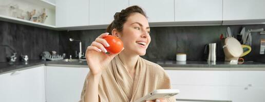 Young woman thinks what to cook, sits in the kitchen with smartphone and tomato in hands, looks aside and smiles, searches recipes on mobile app, orders groceries photo