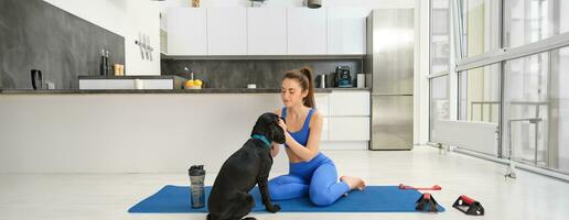 Image of young woman does workout from home, sits on yoga mat in living room and plays with her black dog, puppy distracts girl from doing exercises photo