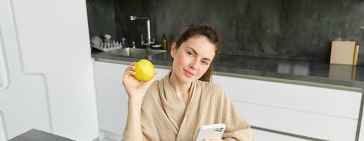 Close up portrait of smiling brunette woman in bathrobe, sits in kitchen at home, uses mobile phone and holds an apple, orders fresh fruits on smartphone app, buys groceries online, looks up recipe photo