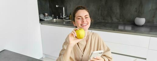 Close up portrait of happy young woman in bathrobe, sitting in the kitchen and using mobile phone, holding an apple, order fruits and vegetables online, using smartphone app for groceries delivery photo