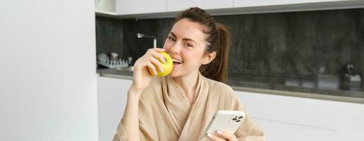 Close up portrait of smiling beautiful woman, sitting in bathrobe, eating apple and looking happy, using smartphone while sitting in the kitchen, order food online on mobile app photo