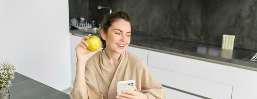 Close up portrait of happy young woman in bathrobe, sitting in the kitchen and using mobile phone, holding an apple, order fruits and vegetables online, using smartphone app for groceries delivery photo