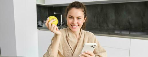 Close up portrait of happy young woman in bathrobe, sitting in the kitchen and using mobile phone, holding an apple, order fruits and vegetables online, using smartphone app for groceries delivery photo