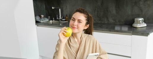 Close up portrait of happy young woman in bathrobe, sitting in the kitchen and using mobile phone, holding an apple, order fruits and vegetables online, using smartphone app for groceries delivery photo