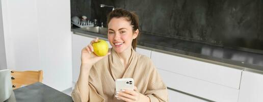 Close up portrait of smiling brunette woman in bathrobe, sits in kitchen at home, uses mobile phone and holds an apple, orders fresh fruits on smartphone app, buys groceries online, looks up recipe photo