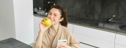 Close up portrait of happy young woman in bathrobe, sitting in the kitchen and using mobile phone, holding an apple, order fruits and vegetables online, using smartphone app for groceries delivery photo