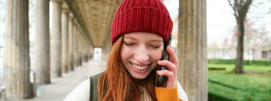 retrato de pelirrojo europeo niña en rojo sombrero, hace un teléfono llamar, camina en ciudad y negociaciones a amigo en teléfono inteligente foto