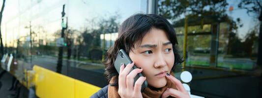 Perplexed asian girl, talks on mobile phone with concerned, thoughtful face, stands near bus stop and frowns, thinking while listening telephone conversation photo