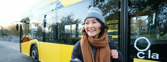 Portrait of girl standing near bus on a stop, waiting for her public transport, schecks schedule on smartphone application, holds mobile phone, wears warm clothes photo