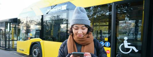 imagen de niña estudiante esperando para público transporte, cheques calendario en teléfono inteligente aplicación, soportes cerca ciudad autobús foto