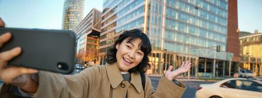 sonriente coreano niña demostración ciudad, tomando selfie en frente de edificios en calle con contento rostro, toma fotos en su teléfono inteligente cámara