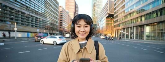 Portrait of young asian girl, student walks in city, listens music in headphones and uses mobile phone on streets photo