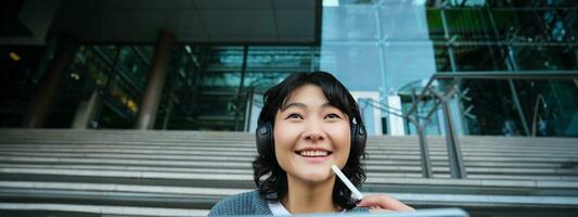 Close up of smiling asian girl feels inspired while draws on digital tablet, makes graphic design project, pencils user interface assignment, sits on stairs near campus photo