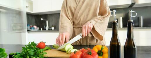 Healthy lifestyle. Young woman in bathrobe preparing food, chopping vegetables, cooking dinner on kitchen counter, standing over white background photo
