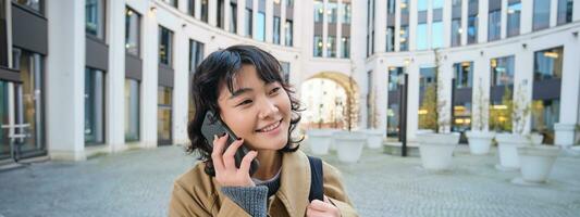celular tecnología. joven coreano mujer negociaciones en móvil teléfono, hace un teléfono llamada en su camino hogar, camina abajo calle, ciudad centro, tiene teléfono conversacion foto