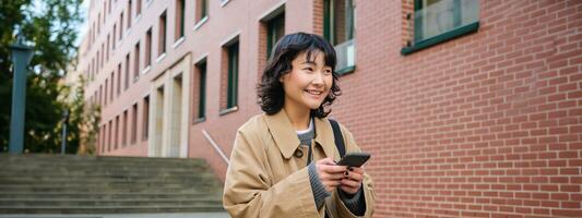 Stylish korean hipster girl, walks in city centre, sends text message, uses location map application on smartphone while walking down the street, wears trench coat photo