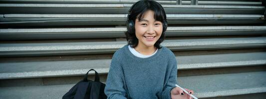 Young asian girl, student in headphones, works on remote, digital artist drawing on tablet with graphic pen, listening music in headphones and sitting on street staircase photo