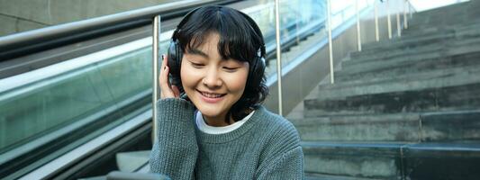 retrato de sonriente coreano niña en auriculares, usos teléfono inteligente y se sienta en escalera en centro comercial, relojes vídeo en móvil teléfono foto