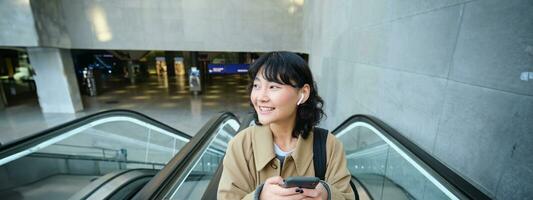 retrato de linda sonriente coreano chica, va arriba escalera mecánica, escucha música en inalámbrico auriculares y usos móvil teléfono, sostiene teléfono inteligente foto