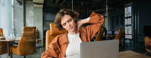 retrato de mujer siente dolor en cuello, se sienta en oficina con computadora portátil, tiene tensión en músculos después trabajando todas día foto