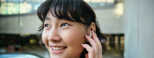 Close up portrait of smiling korean girl in headphones, listens music in wireless earphones, enjoys travelling around town with her favorite songs playlist photo