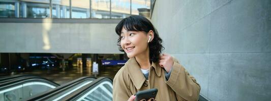 Young brunette woman commutes, goes somewhere in city, stands on escalator and uses mobile phone, holds smartphone and smiles, listens music in wireless headphones photo