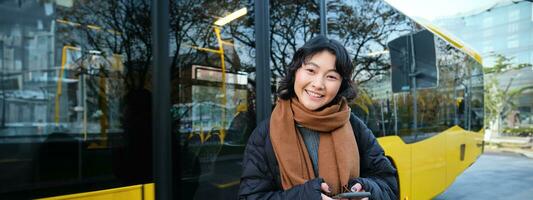 Portrait of korean girl buying ticket for public transport online, using mobile application on bus stop, wearing winter clothes photo