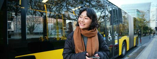 Portrait of korean girl buying ticket for public transport online, using mobile application on bus stop, wearing winter clothes photo