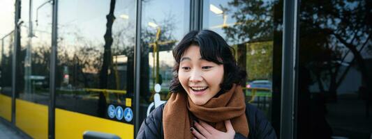 Portrait of cheerful asian girl talks on mobile phone, video chats, looks amazed at smartphone camera, stands on bus stop photo